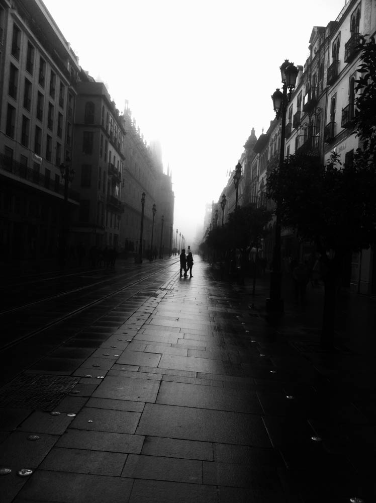 Two figures walk down a wet, empty street in Seville, its tall buildings dissolving into fog.