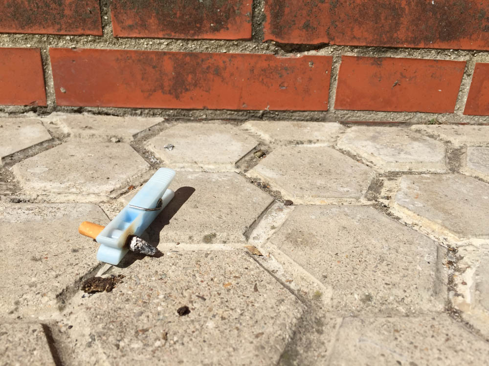 Blue plastic tweezers gripping a cigarette butt on hexagonal paving beside a brick wall.