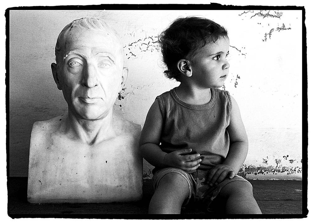A small boy sits beside a white stone bust, both turned away from the camera's gaze.