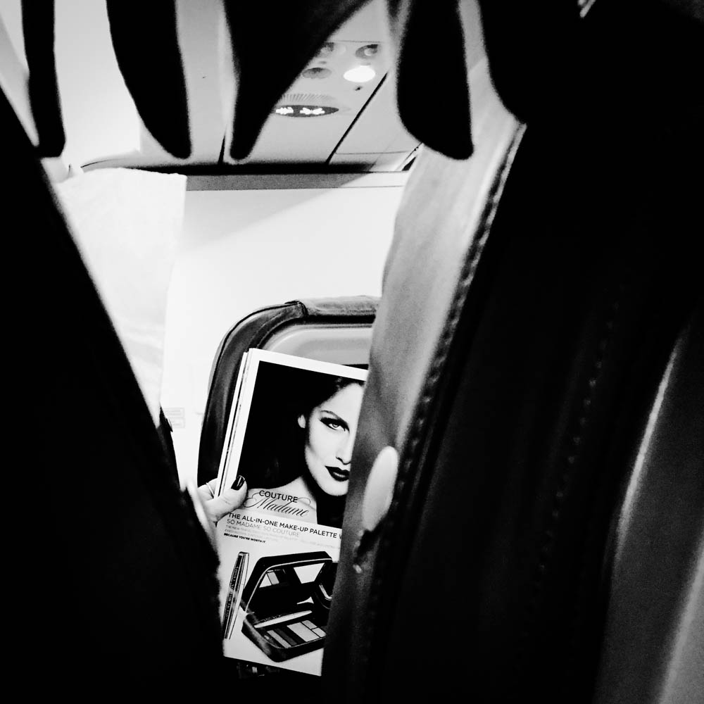 Through a gap between aeroplane seats, a woman's face looks back from a magazine page.