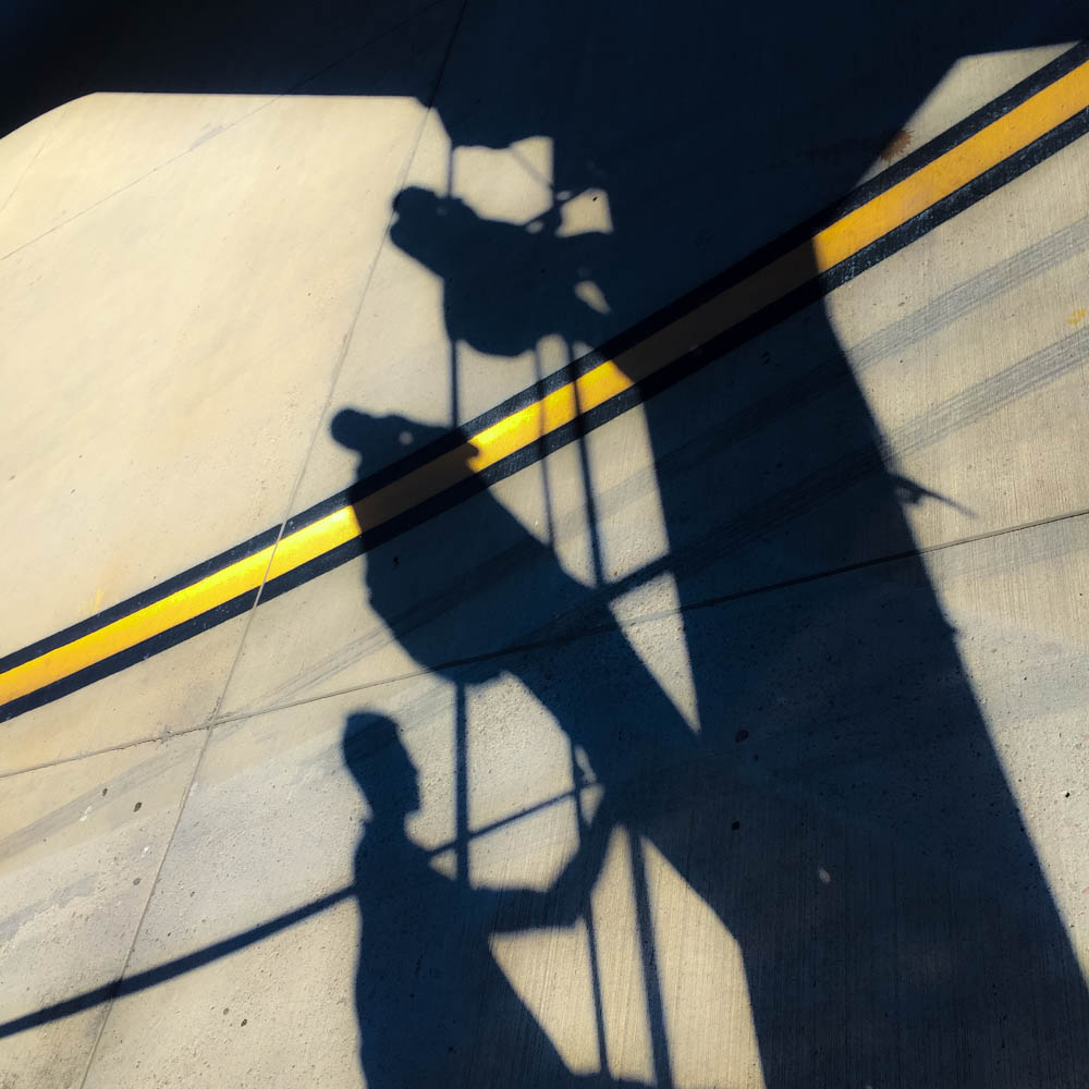 Shadows of people climbing aircraft boarding stairs, cast across pale ground crossed by a yellow line.